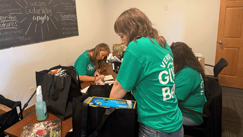 Volunteers in green shirts pack bags of supplies around a conference table in a community organization workspace.